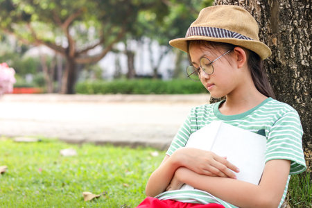 Primary girl sitting reading a book until sleeping, lying under the treeの写真素材