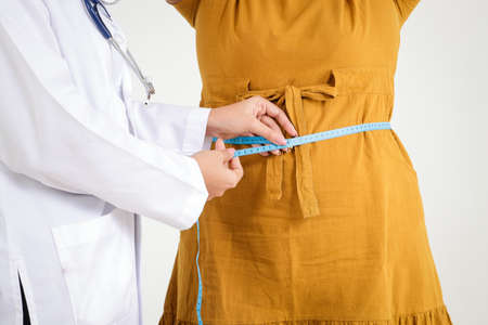 Female doctor wearing white uniform Hold a tape measure around the waist of a obese woman wearing a yellow dress has health and obesity problems. Concept of patient treatment, weight loss.の写真素材