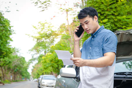 An Asian man has crashed into a car accident. Park your car on the road, hold paperwork, and call the car insurance company. Concept of insurance. Copy spaceの写真素材