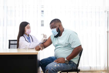 An Asian female doctor wearing a mask and gloves is vaccinating an African-American manの写真素材