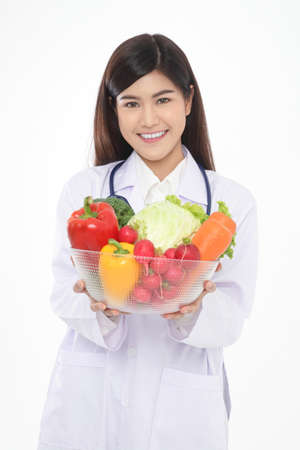 Beautiful young Asian female doctor has a beautiful smile. Holding a bowl of fresh organic vegetables Food for good health. Prescription food nutrition concept, healthy eating. White background.の写真素材