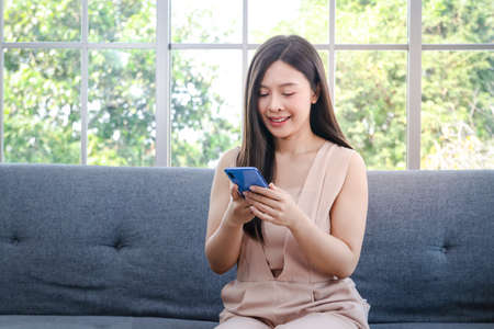 A beautiful smiling young woman holding a smartphone Sit on the sofa in the house. Concept of rest on vacation.の写真素材