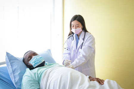 An Asian female doctor wears injection gloves to an African-American male patient lying in bed. Concept of hospital medical serviceの写真素材
