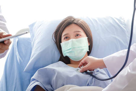 Female doctor holding a stethoscope, doctor examining the heart, Asian woman patient Wear a blue mask and lie in a hospital bed. Concept of medical service, health insurance. Copy spaceの写真素材