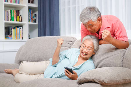 Asian elderly couple in living room, happy, joyful, cheerful holding a black smart phone. View online media on the Internet. Retirement life concept, social distancingの写真素材