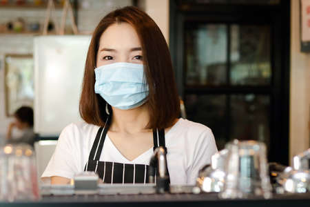 A beautiful Asian female employee wearing a mask and an apron. standing at the coffee counter Ready to serve customers during the virus spread. entrepreneurial concept for small business.の写真素材