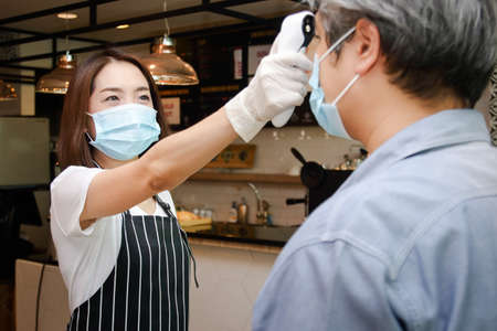 A beautiful Asian female employee wearing a mask and wearing gloves Measure the temperature on the forehead of customers who come to use the service. to prevent coronavirusの写真素材