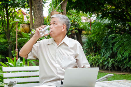 Asian elderly man He uses his laptop to work in the garden of his residence. He was holding a glass and was drinking plain water. concept of health care for seniors in retirement ageの写真素材