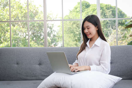 asian girl sitting on the sofa in the living room at home Working on a laptop computer. Work from home concept, social distancing During the  pandemicの写真素材