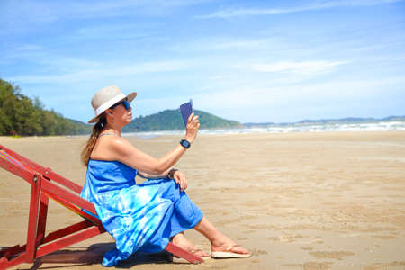 Stylish Asian woman wearing a long blue dress. Sunbathing by the sea holding a smartphone to take pictures of beautiful natural scenery Travel to the sea in Thailand, tourismの写真素材