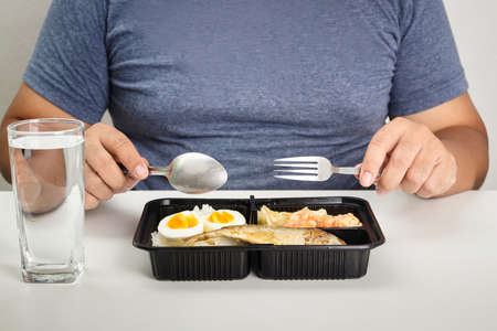 Fat man eating a box of food on a white table. Concept of home quarantine for patients asymptomatic, mild symptoms while waiting for bedの写真素材