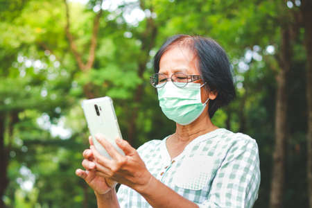 Asian elderly women wearing green masks standing in the garden holding a smartphone Talk online via...