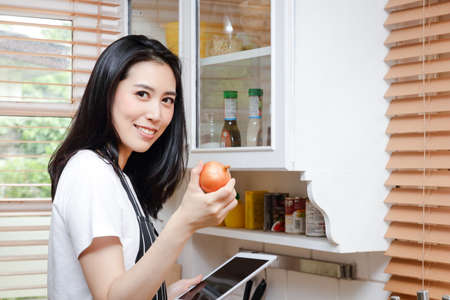 Beautiful Asian woman practicing cooking in the kitchen at home Hold your tablet to watch online cooking videos. Concept of living during the pandemic.  social distancingの写真素材