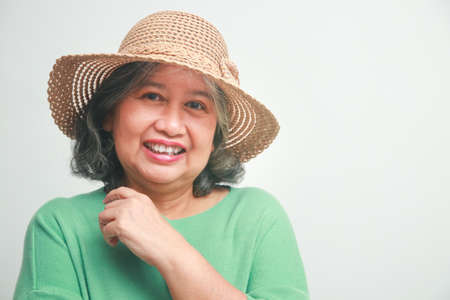 Asian elderly woman wearing a green shirt and a hat She smiles happily, getting dressed for a trip. Standing on a white background. travel concept Elderly people are happy in retirement.の写真素材