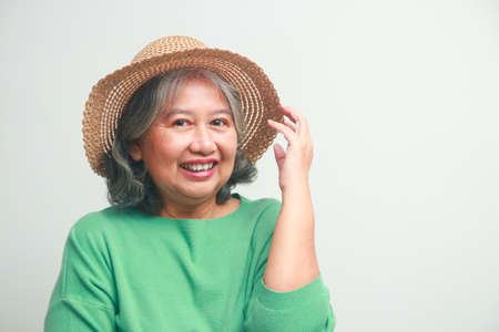 Asian elderly woman wearing a green shirt and a hat She smiles happily, getting dressed for a trip. Standing on a white background. travel concept Elderly people are happy in retirement.の写真素材