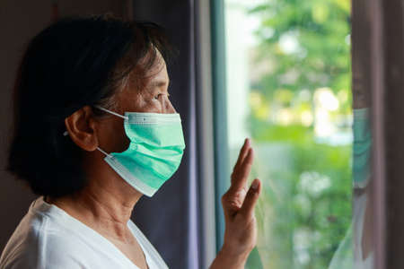 Asian elderly woman wearing masks living at home Standing and looking out at the front of the house, bored of having to detain himself. home isolation, social distancingの写真素材