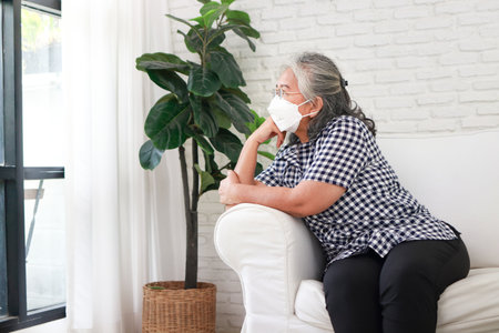 Asian elderly women wearing masks She sat lonely on the sofa in the house. Taking care of yourself at home during the pandemic.の写真素材