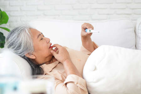 Elderly Asian woman lying sick on the sofa in the house Take medication for fever as prescribed by the doctor. and use a digital thermometer to check the temperature. treating yourself at homeの写真素材