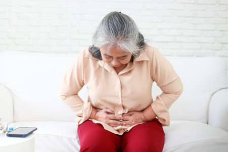 Asian elderly woman with severe abdominal pain sitting on the sofa in the house Use your hands to press against your stomach to reduce the pain. Gastrointestinal problems in the elderlyの写真素材