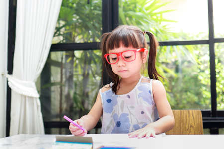 cute little asian girl Wearing red glasses, standing on the table at home drawing drawings. educational concepts, develop children's learningの写真素材
