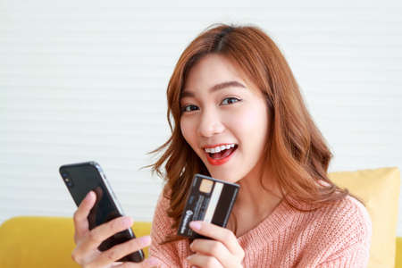 beautiful asian woman live in an apartment holding a black smartphone and credit card to order online. Concept of using modern technology in daily life. Work from home.の写真素材