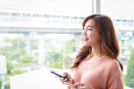 beautiful asian woman Lives in an apartment, standing at a glass window. holding a black smartphone...