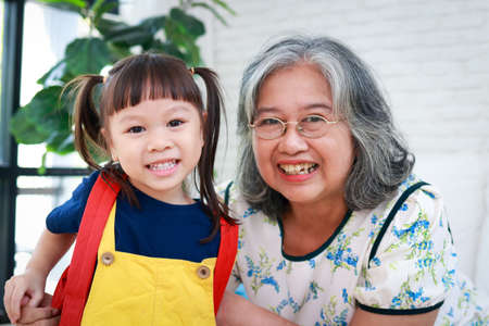 elderly grandmother with cute little granddaughter live at home together. Grandma dressed her granddaughter for school in the morning. educational concepts of kindergarten childrenの写真素材