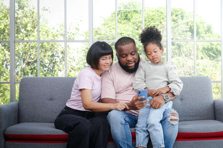 black family parents and daughter sitting on the sofa in the living room They make video calls online on smartphones. Enjoy life at home. Family concept, use of technology to communicate.の写真素材