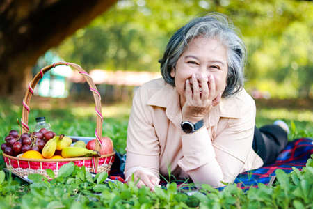 Asian elderly woman picnicking in the park Lying on the grass with a basket of fruit next to it. She is enjoying her vacation. The concept of living in retirement to be happy, eating healthy food.の写真素材