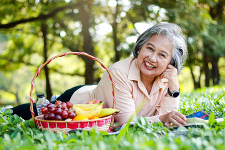 Asian elderly woman picnicking in the park Lying on the grass with a basket of fruit next to it. She is enjoying her vacation. The concept of living in retirement to be happy, eating healthy food.の写真素材