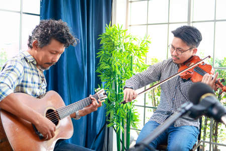 two asian men playing the violin classical instrument and acoustic guitar together in the music practice room The couple enjoyed playing acoustic music. Concept of playing live music and bandの写真素材