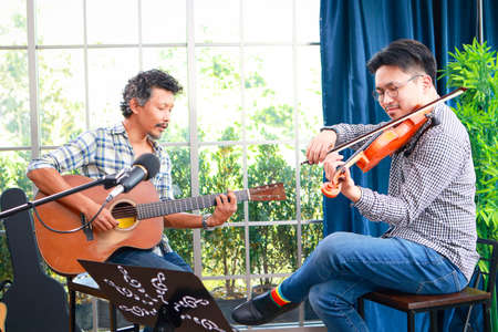 two asian men playing the violin classical instrument and acoustic guitar together in the music practice room The couple enjoyed playing acoustic music. Concept of playing live music and bandの写真素材