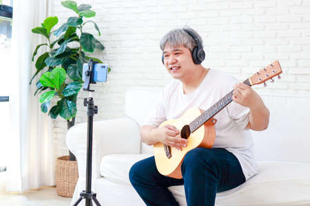 Asian elderly man Play music at home in the living room. Live Mobile broadcasting through applications on smartphones. The concept of playing music online. Modern communication. new lifestyleの写真素材