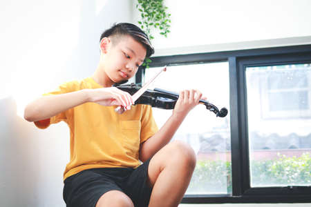 An Asian boy sits at home in the morning playing music. He was happily playing the violin of a...
