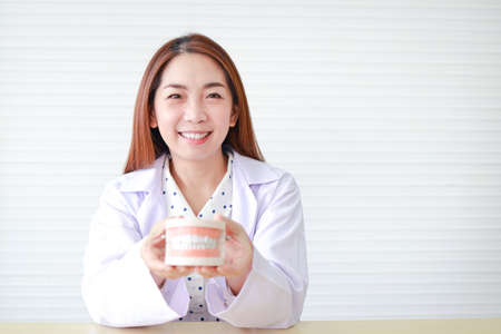 Asian female dentist Sitting in the office, she holds a denture mockup to demonstrate how to keep her teeth clean. concept of medical services in a hospital.の写真素材