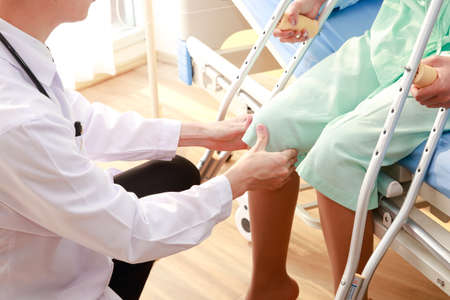 Asian male doctor examining knee of female patient sitting on nursing bed She holds a cane to help her walk. Concept of osteoarthritis, knee pain, osteoarthritis, knee inflammation. Physical therapyの写真素材