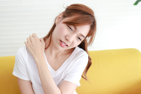 A beautiful Asian woman with pain in her right shoulder sits on the sofa in the living room. The concept of shoulder pain in adults. Office Syndrome. Health care.の写真素材