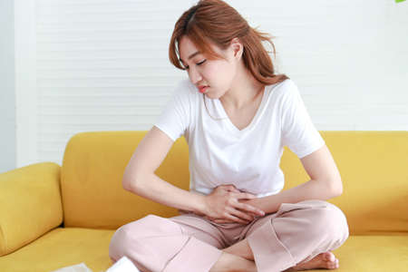 Beautiful Asian woman with lower abdominal pain. She sits on the sofa at home. Concept of treatment of infection, inflammation, digestive system, reproductive system in women. health careの写真素材