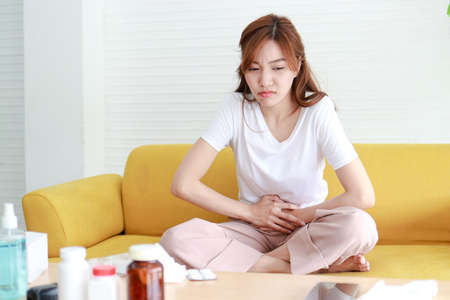 Beautiful Asian woman with lower abdominal pain. She sits on the sofa at home. Concept of treatment of infection, inflammation, digestive system, reproductive system in women. health careの写真素材