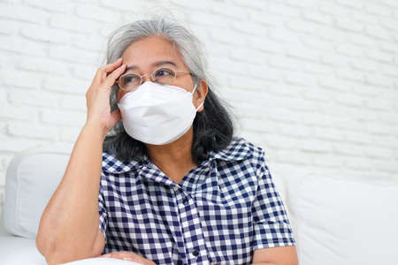 An elderly Asian woman wearing a mask is sitting on the sofa in her home.の写真素材