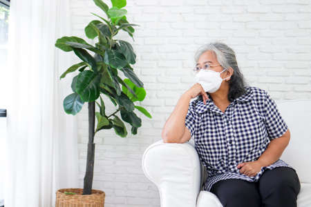 An elderly Asian woman wearing a mask is sitting on the sofa in her home. She was worried about health problems. There is no one to take care of. The concept of health care for the elderly.の写真素材