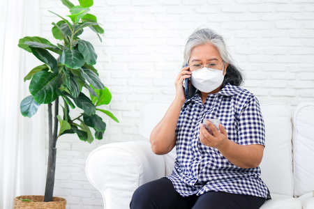 Online Medicine Concept. An elderly Asian woman has a fever. Holding a black smartphone, talking to a doctor online, asking how to use medicine. Social distancing to prevent viral infectionの写真素材