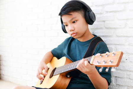 Asian boy wearing headphones with black music Sit and play guitar in the house. music learning concept, music proficiency training. copy spaceの写真素材