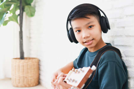 Asian boy wearing headphones with black music Sit and play guitar in the house. music learning concept, music proficiency trainingの写真素材