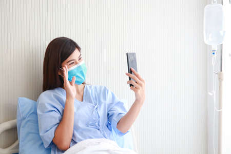 Asian female patient wearing mask in hospital patient room She holds a smartphone online talking to her family.の写真素材