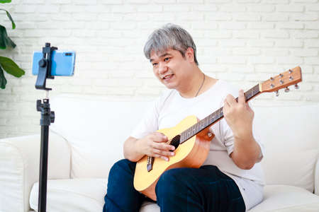 Asian white haired man playing music at home enjoying the music. Learn music online through your smartphone. leisure activities. modern communication new lifestyleの写真素材