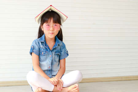 Cute little Asian girl having fun, she wears pink glasses and puts a book on her head. Sit on the wooden floor in the house. education concept learning for kids.の写真素材