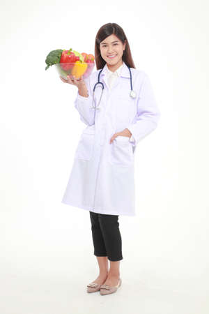 Beautiful young Asian female doctor has a beautiful smile. Holding a bowl of fresh organic vegetables Food for good health. Prescription food nutrition concept, healthy eating. White background.の写真素材