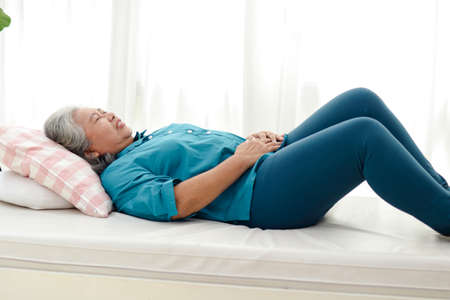 An elderly Asian woman lies in bed with acute abdominal pain at home. Concept of problems of...