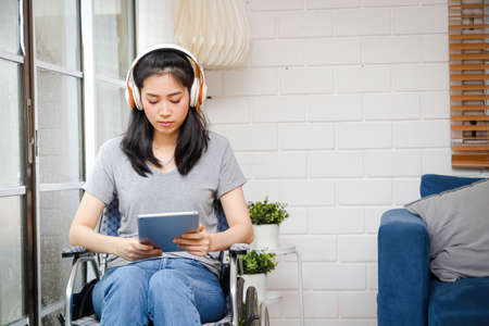Asian women can't walk. wearing headphones playing tablet sitting in a wheelchair at home Happy with life.の写真素材